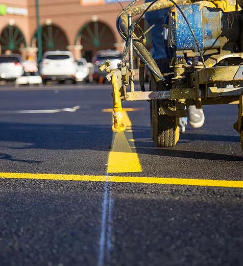 A line striping machine applying yellow paint to a freshly paved parking lot with vehicles in the background.